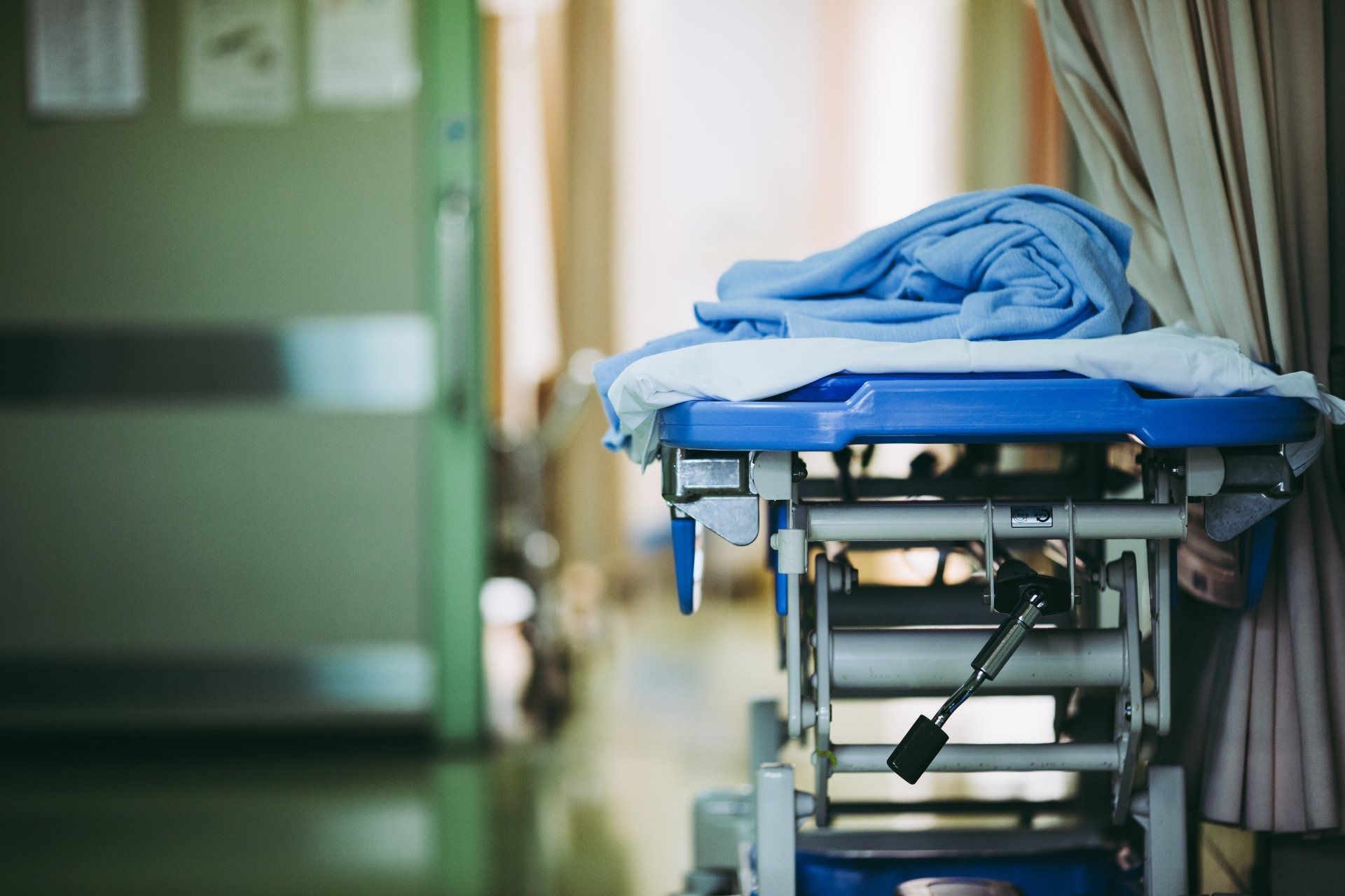 Hospital gurney with blue blanket in a hallway; green door on the left, beige curtain on the right.