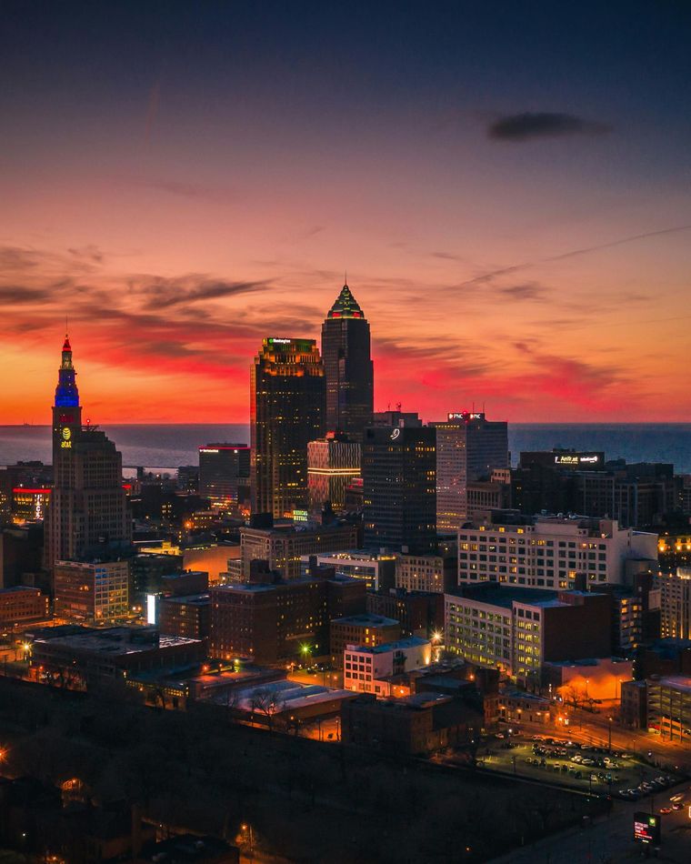 Cleveland skyline at sunset, orange and blue hues in the sky, city lights illuminate buildings.