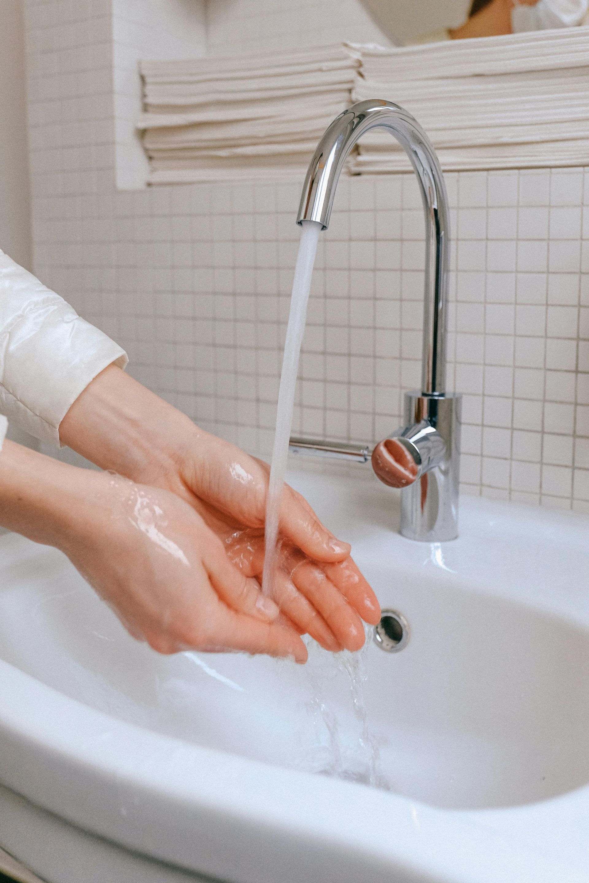 Hands under running water in a white sink, chrome faucet, white tiled wall.