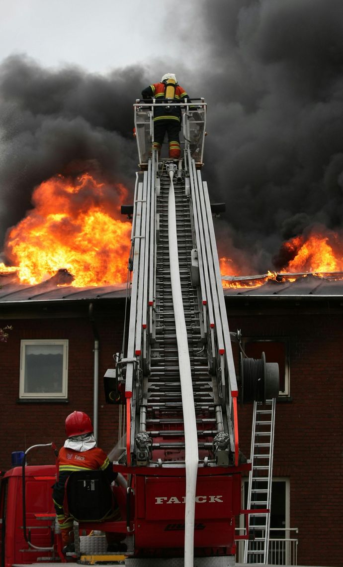 Firefighters on a ladder truck spraying water on a burning building; thick smoke and flames visible.