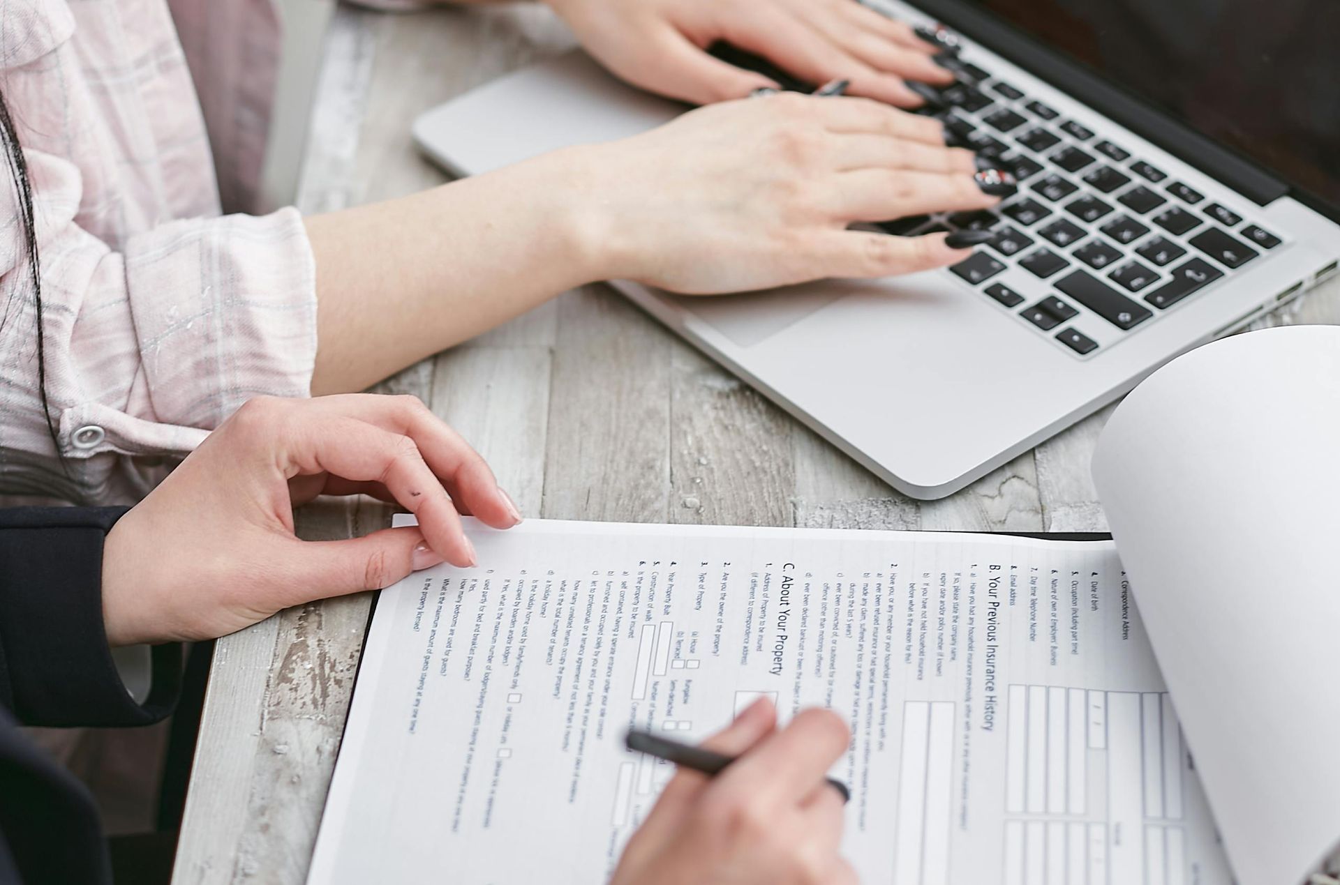 Two people working on a laptop and documents at a table. One types, the other holds a pen.