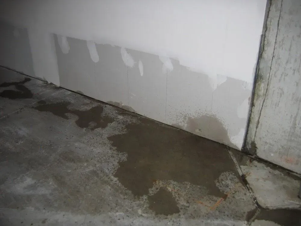 Wet concrete floor with water pooling near a white wall with damp patches.