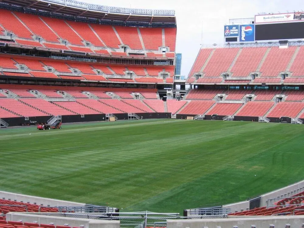 Empty football stadium with brown seating, green field, and cloudy sky.