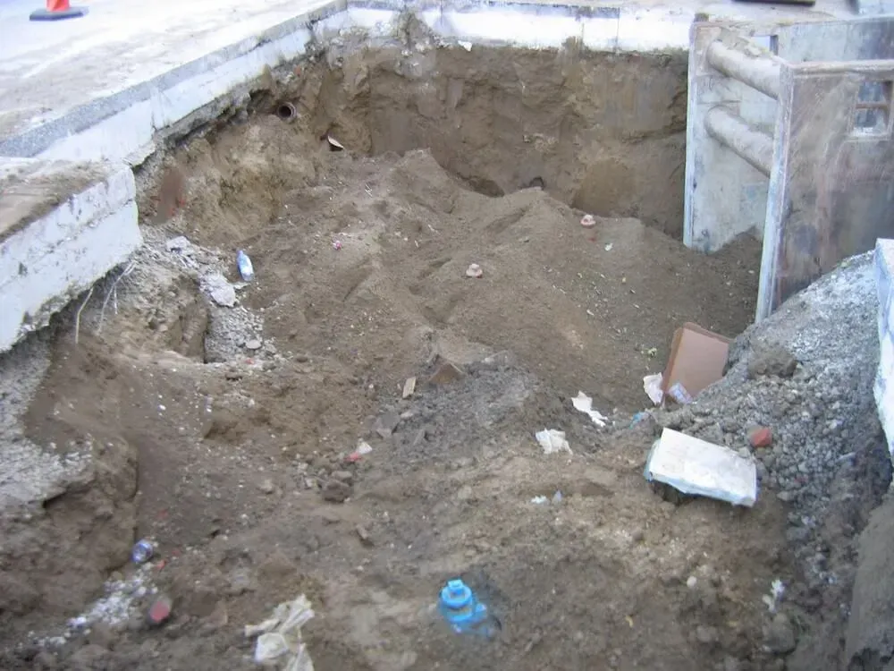 A deep excavation with loose dirt and construction debris along a sidewalk.