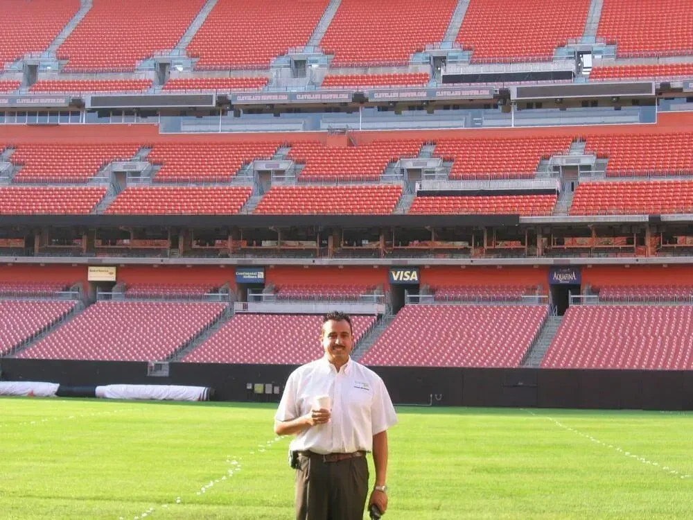 Man stands on a green field in an empty stadium, rows of red seats in the background.