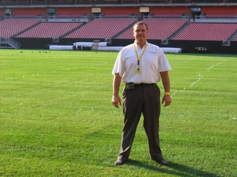 Man in tan shirt and brown pants stands on a green football field, stadium seating in the background.
