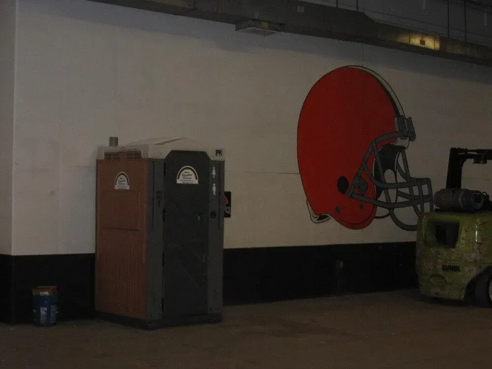 A Cleveland Browns helmet mural above two portable toilets and a forklift in a concrete room.