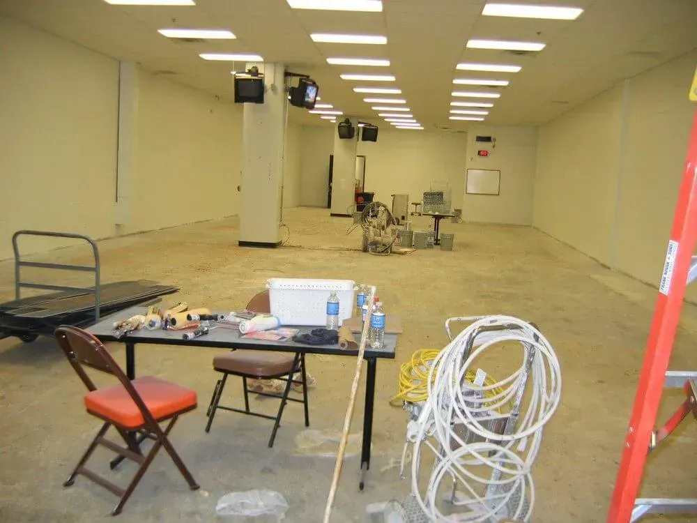 Empty room with work table and tools in the foreground. Overhead lighting, camera monitors on walls. Red ladder.