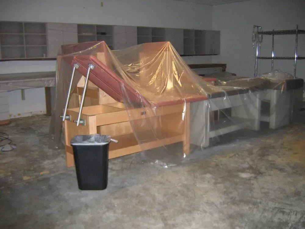 A medical exam table and cabinet covered in plastic sheeting in a room with a concrete floor.