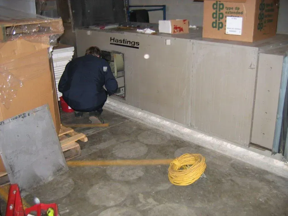 Person crouched, working on a Hastings HVAC unit in a basement setting. Yellow electrical cord on floor.
