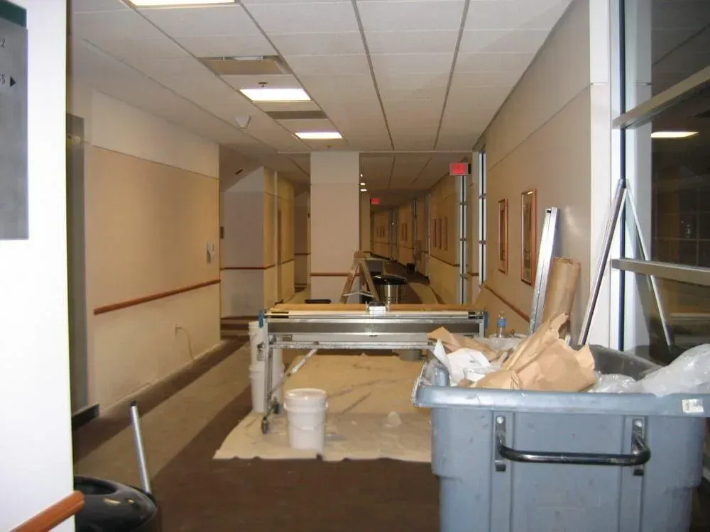 Long hallway undergoing renovation; tools, supplies, and drop cloths present. Beige walls, fluorescent lighting.