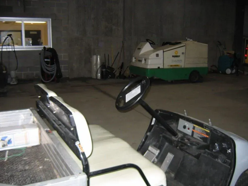 A golf cart inside a warehouse. A cleaning machine is in the background. Fluorescent lights and a small window are visible.
