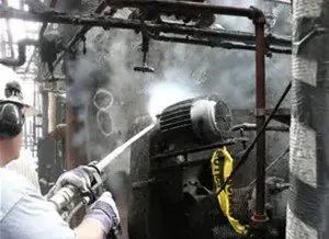 Man spraying equipment with a high-pressure cleaner, industrial setting, smoke present.