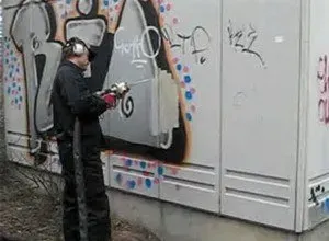 A person removing graffiti from a white electrical box with a power washer outdoors.