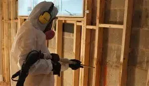 Person in hazmat suit sprays an unfinished interior wall.
