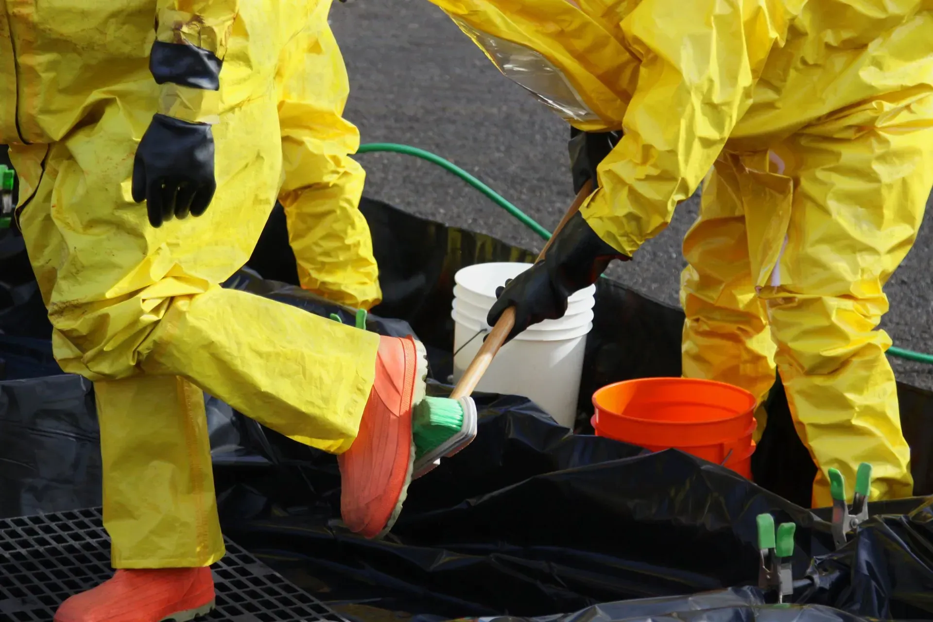 Two workers in yellow hazmat suits cleaning up spill, near buckets and black tarp.
