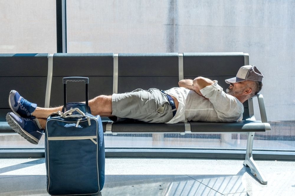 A man is laying on a bench with his feet up next to a suitcase.