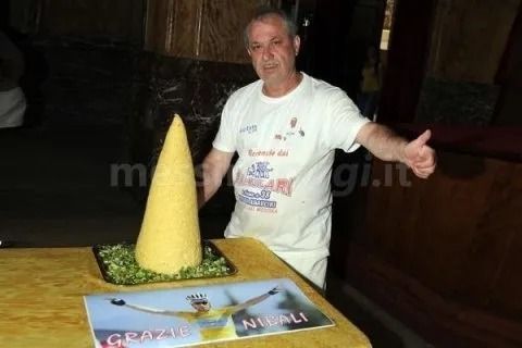 Uomo in camicia bianca con i pollici alzati accanto a un grande piatto di cibo a forma di cono su un tavolo