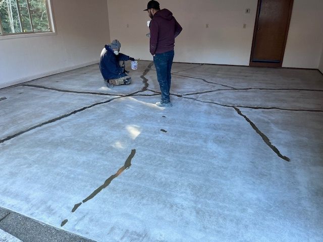 Two people filling cracks in a concrete floor indoors. Gray floor, tan walls, one kneeling, one standing.