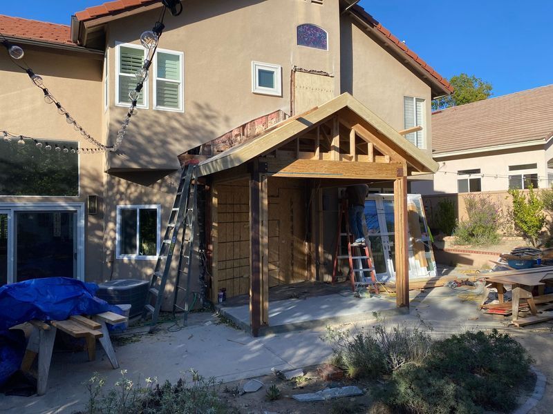 Backyard house addition under construction with wooden porch framing and ladders beside stucco home