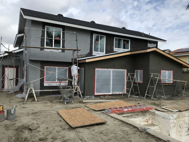 Two-story house under construction with scaffolding, workers, and unfinished siding and dirt yard
