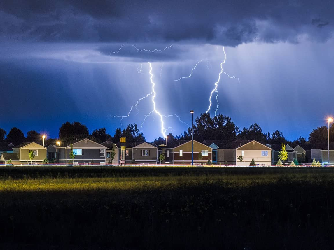 Lightning strikes over a suburban neighborhood at night; dark clouds and bright flashes of light.