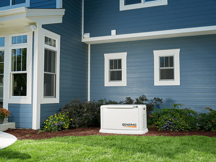 A Generac home backup generator sits on grass next to a blue house with white trim and windows.