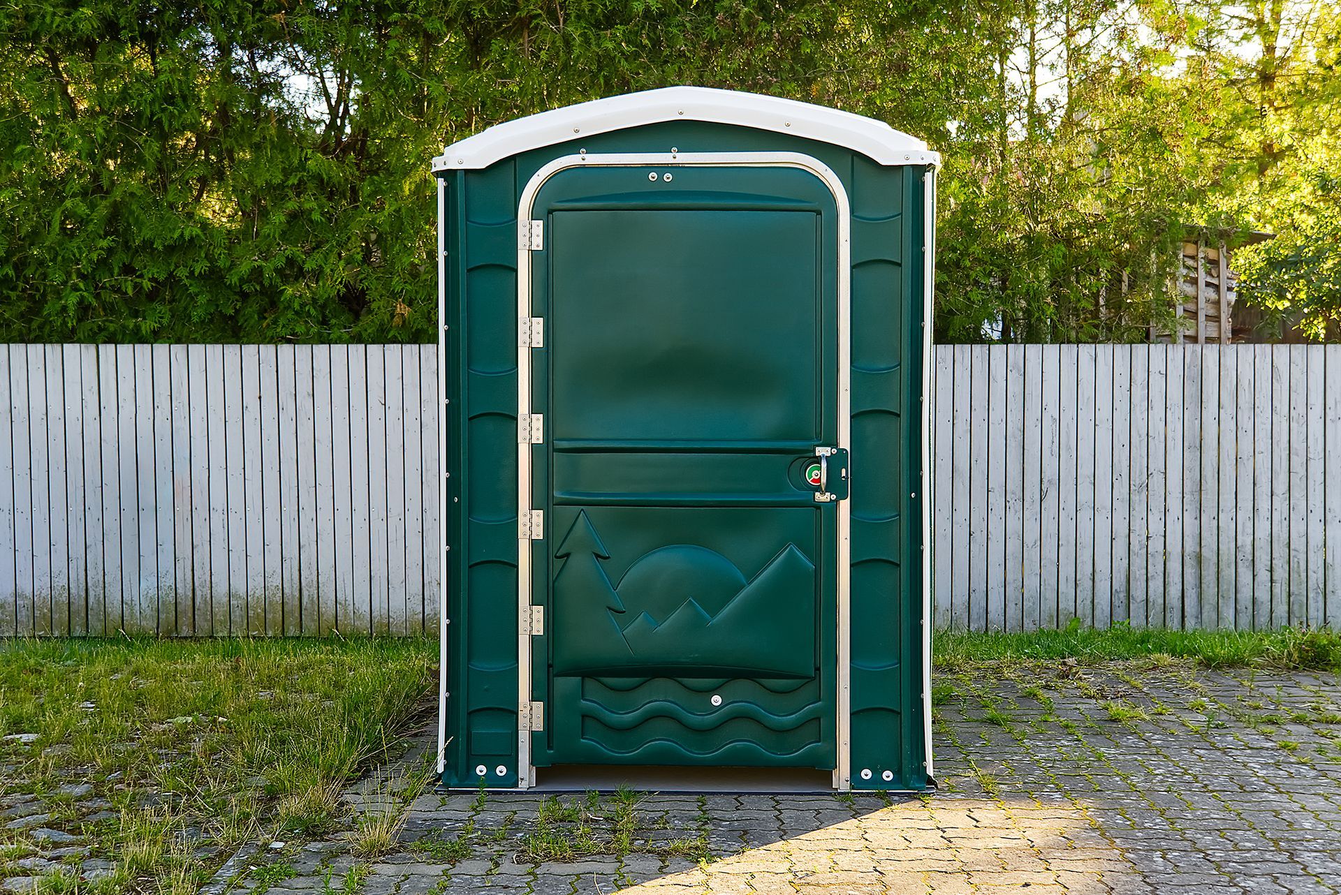 A green portable toilet is sitting in front of a wooden fence