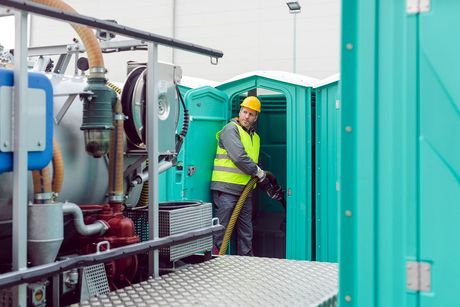 A man in a yellow hard hat is standing in front of a green portable toilet.