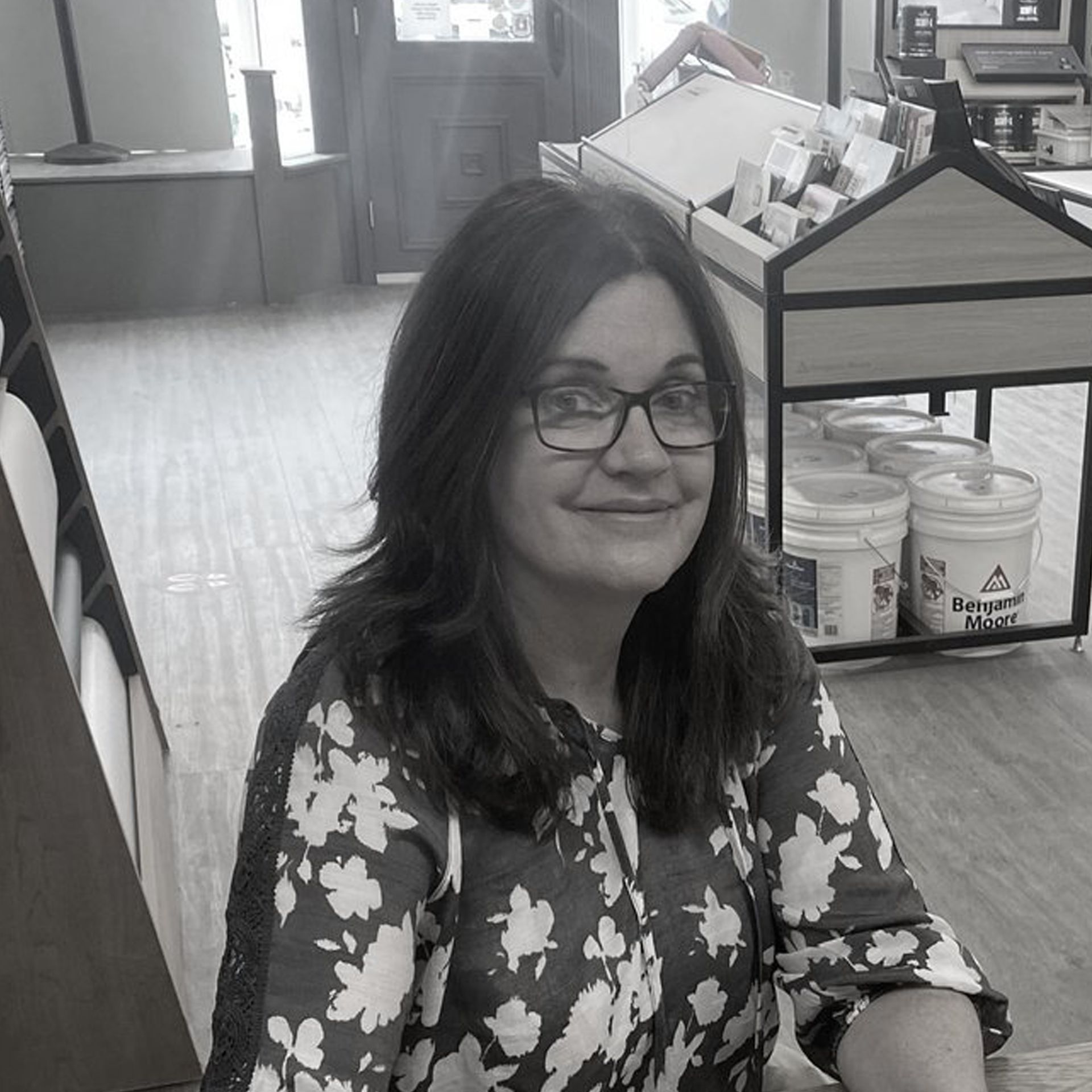 A woman wearing glasses and a floral shirt is smiling in a black and white photo.