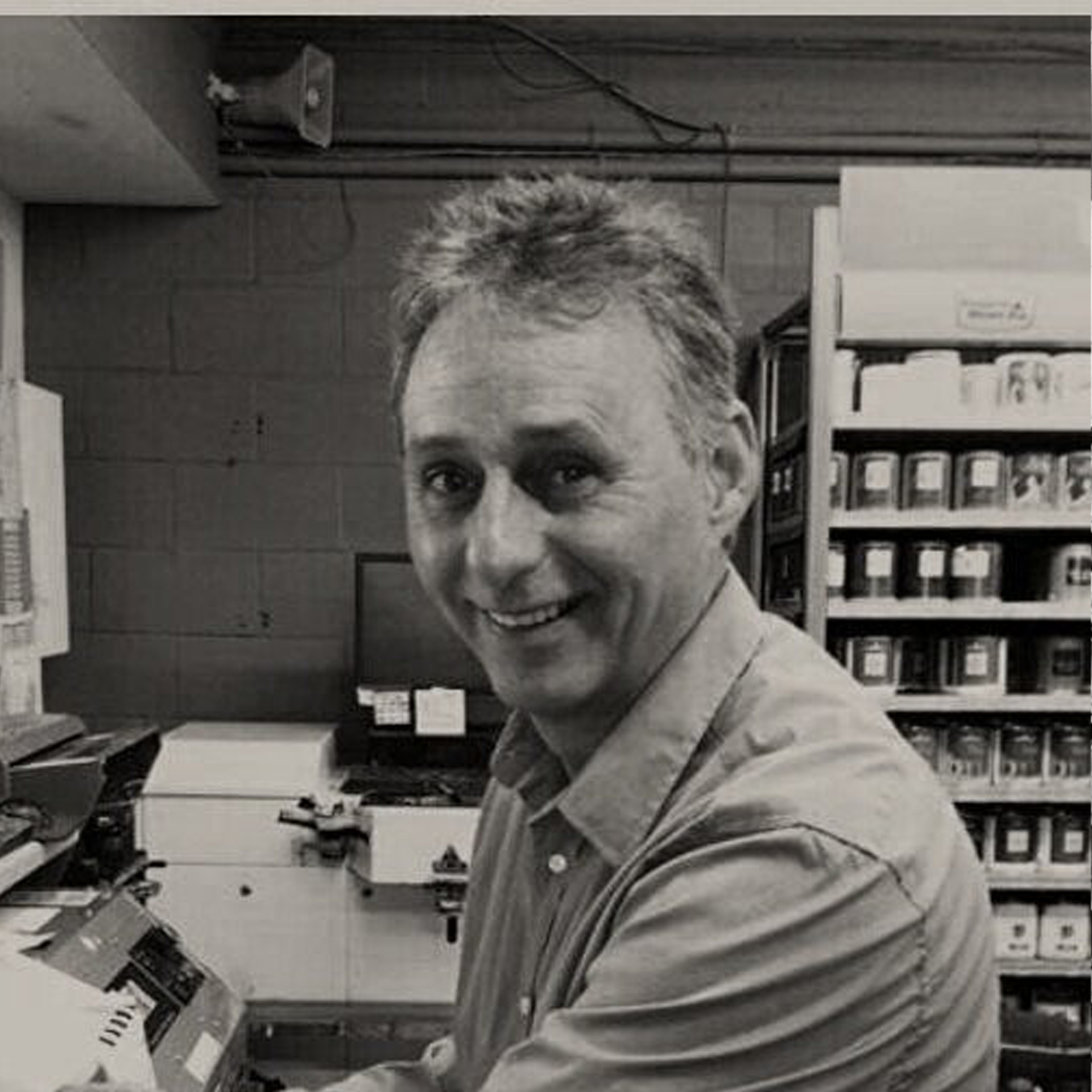 A man is smiling in a black and white photo while sitting at a counter in a store.