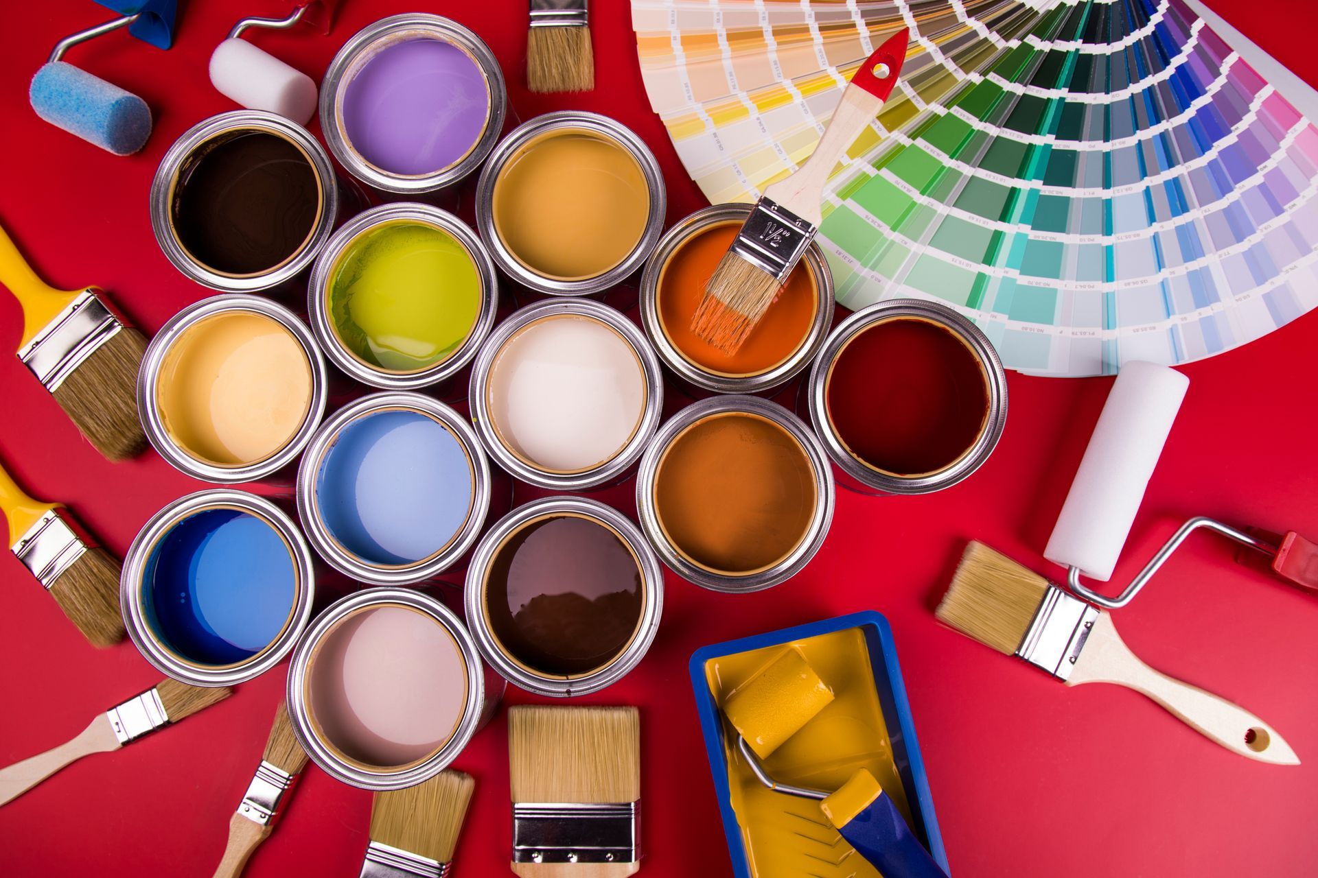 Open paint cans with colors arranged on a red table beside brushes, rollers, and color swatches