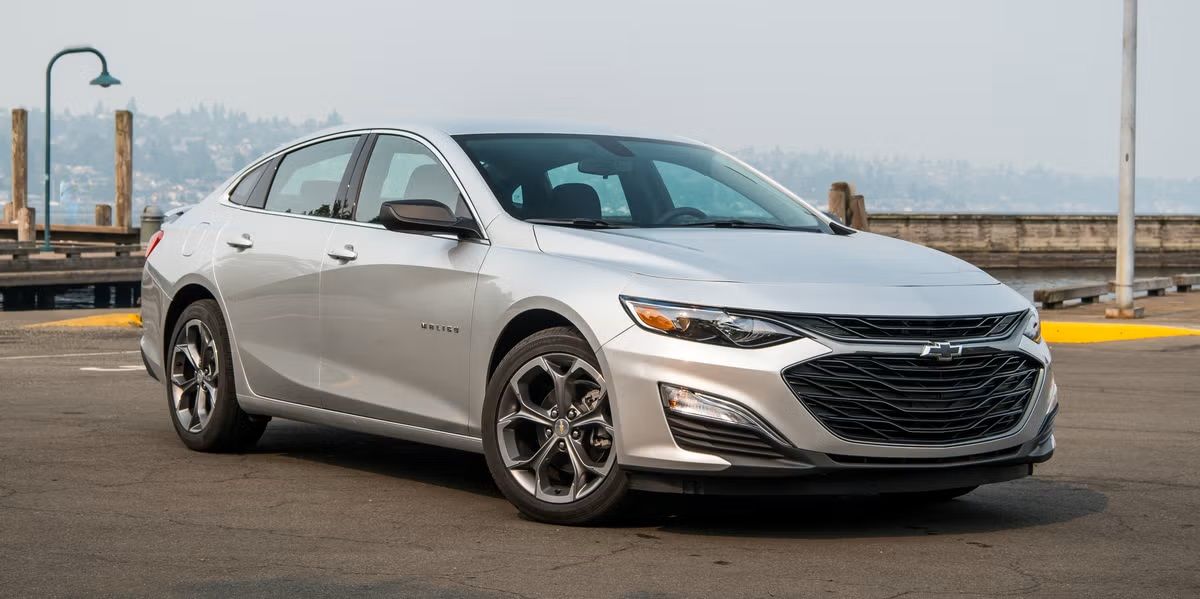 Silver Chevrolet Malibu sedan parked on a pier with water and hills in the background.