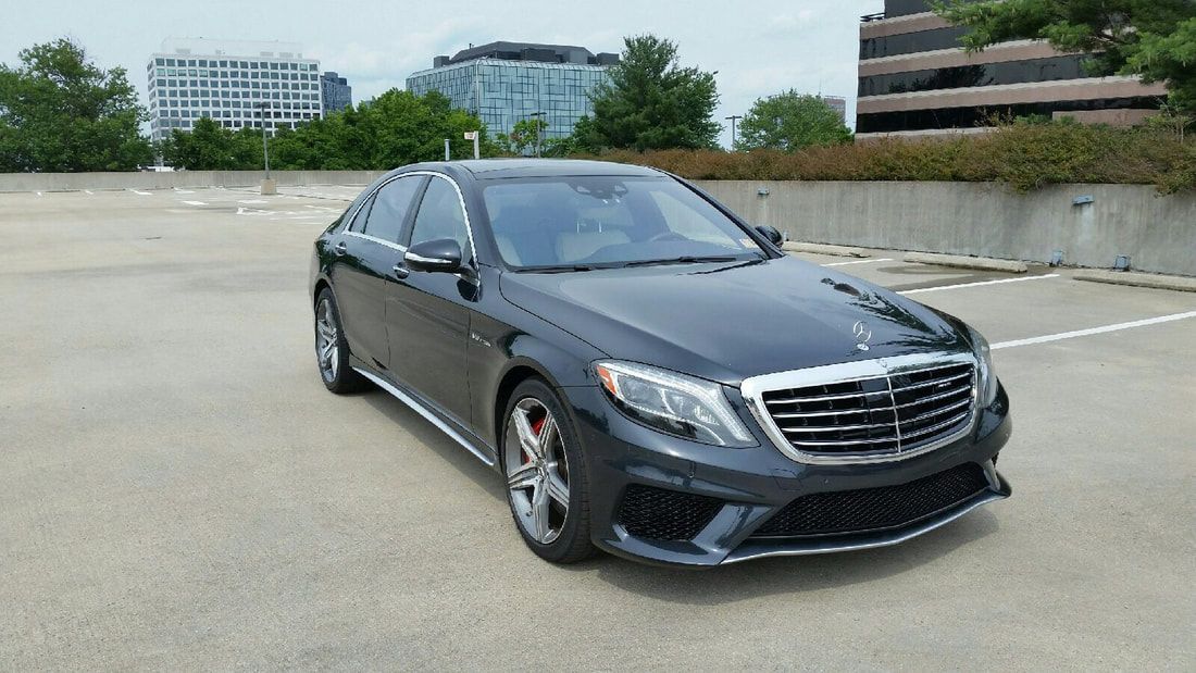 Dark gray Mercedes-Benz S-Class sedan parked in a parking lot, with city buildings in the background.