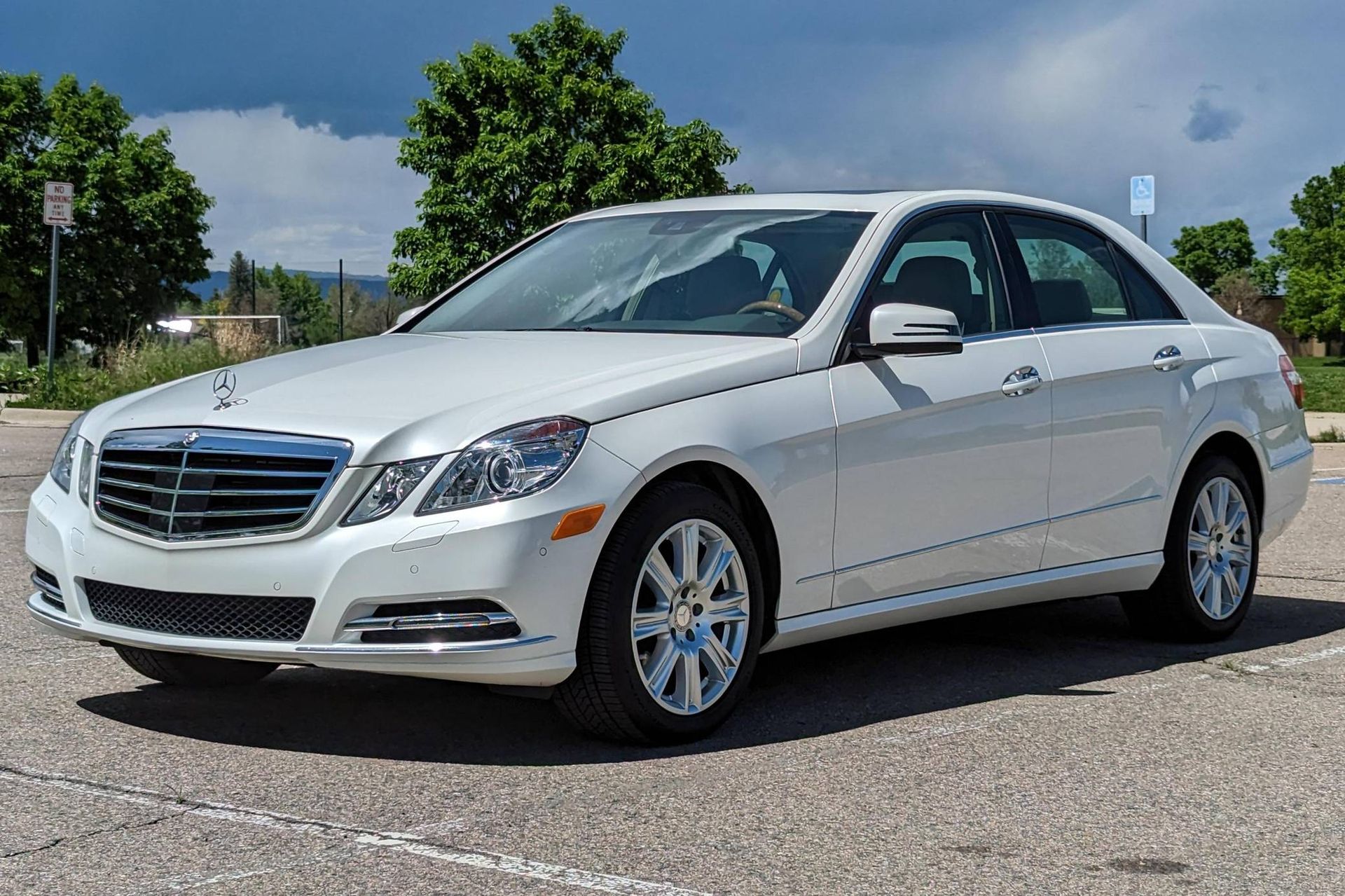 White Mercedes-Benz sedan parked on asphalt in a sunny outdoor setting with trees and a cloudy sky.