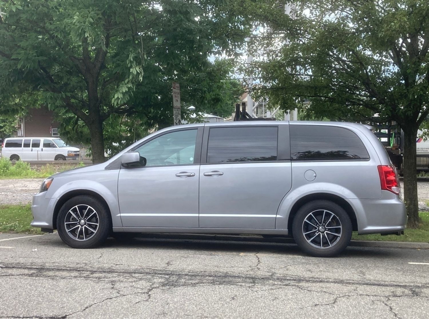 Silver minivan parked on a paved street next to a grassy area with trees and a building in the background.