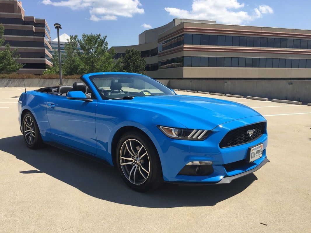 Blue convertible Mustang parked in a parking lot, sunny day, buildings in the background.
