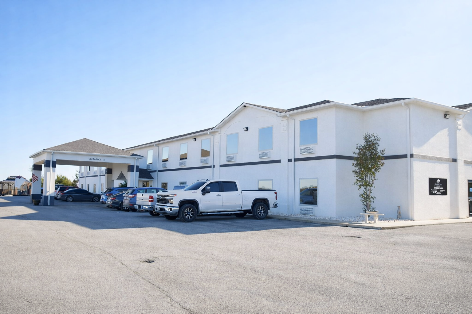 A white, two-story motel building with a covered entrance and a parking lot filled with various cars on a sunny day.