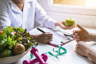 A nutritionist in a lab coat holds a green apple while consulting with a client, with fresh produce and weights on a desk.
