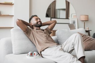 Man relaxing on a couch, hands behind head, smiling. Beige interior, headphones nearby.