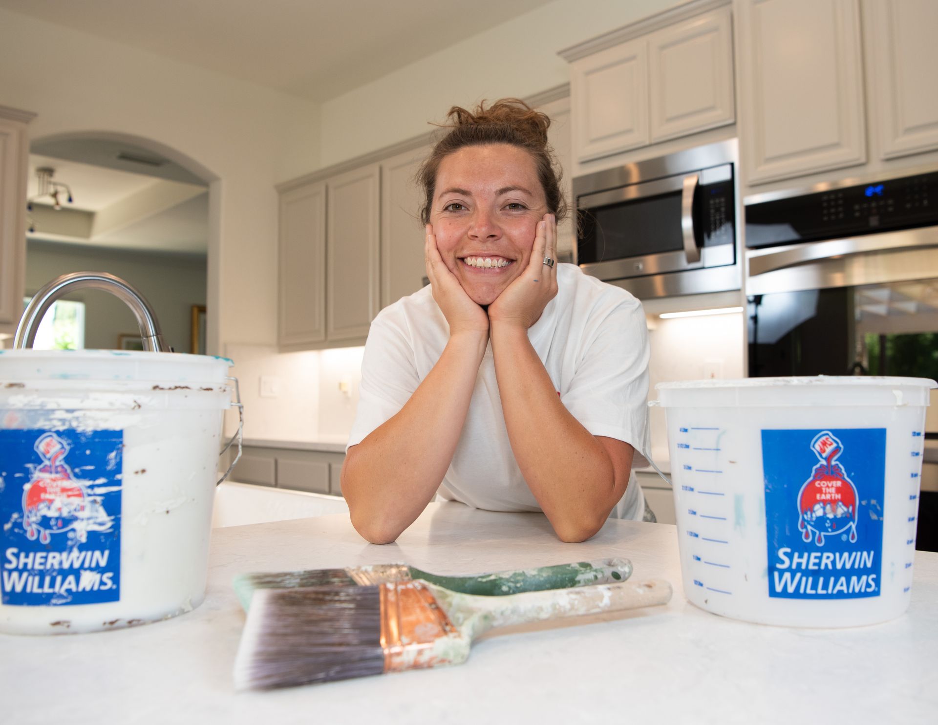 A woman is sitting at a counter with two buckets of sherwin williams paint and a brush.