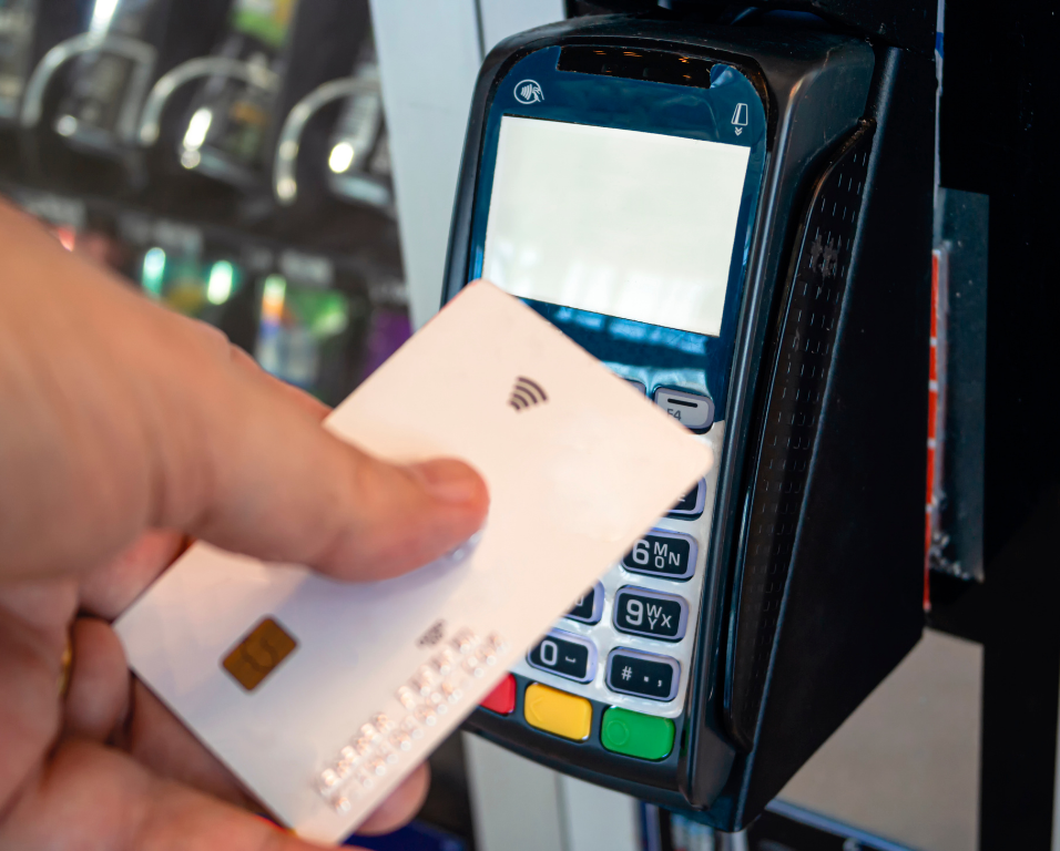 A hand holds a white contactless payment card near a card reader mounted on a vending machine.