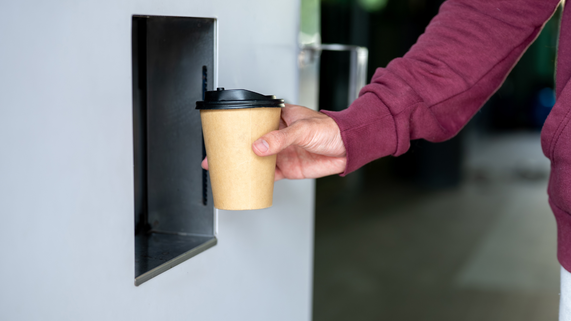 A person in a maroon sweater holding a beige paper coffee cup with a black lid near a gray dispenser.