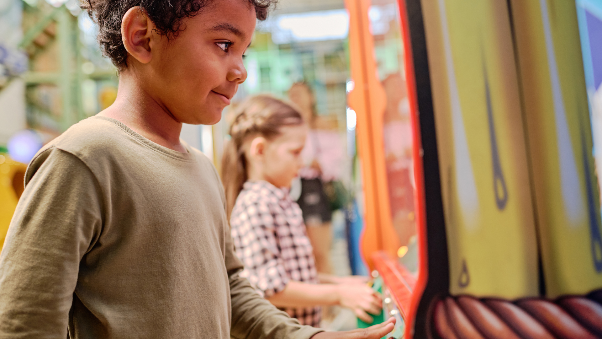 A boy and a girl stand side-by-side playing at an interactive arcade game in a brightly lit indoor space.