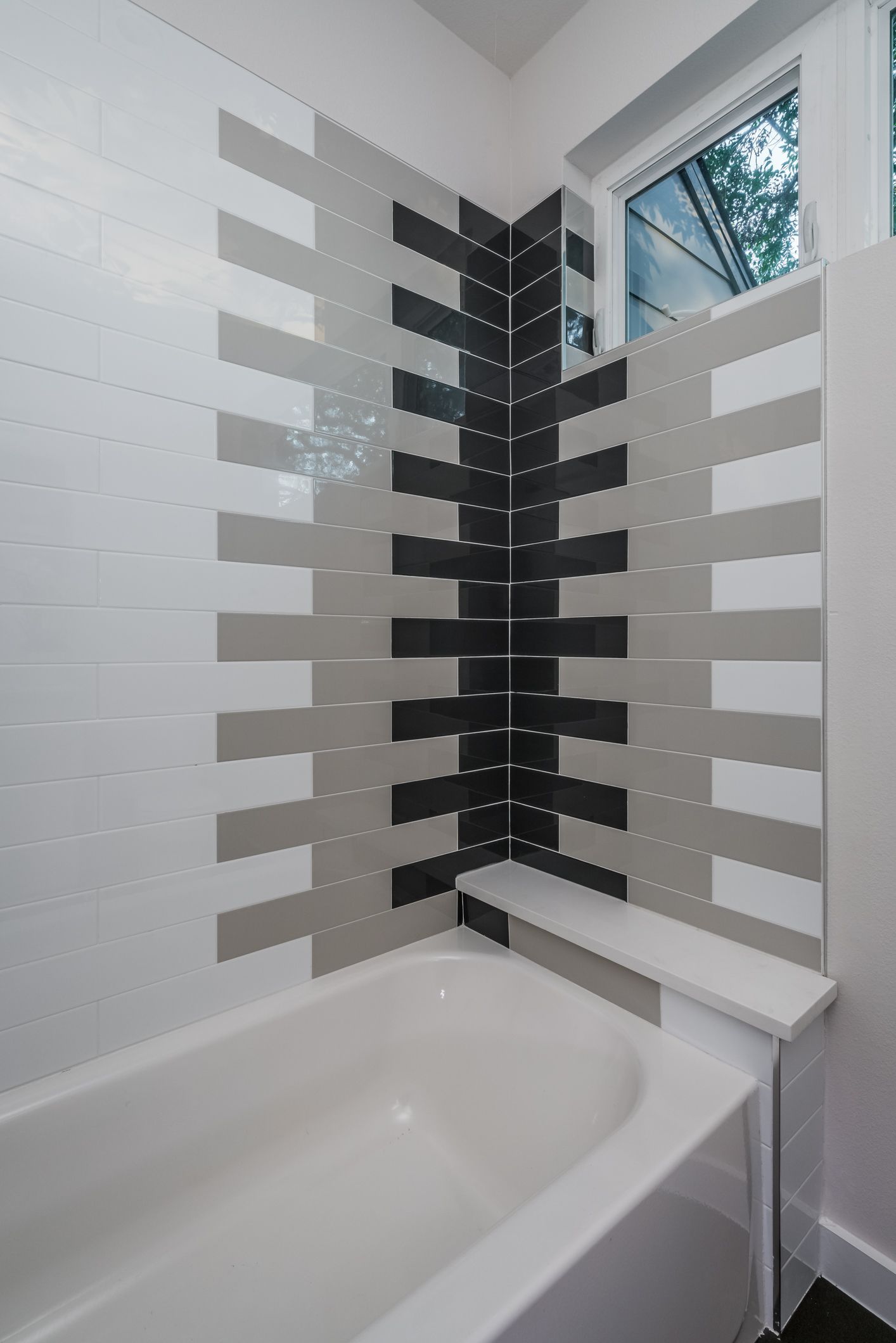 A bathtub with tiled walls featuring a geometric pattern of white, gray, and black rectangular tiles in a corner layout.