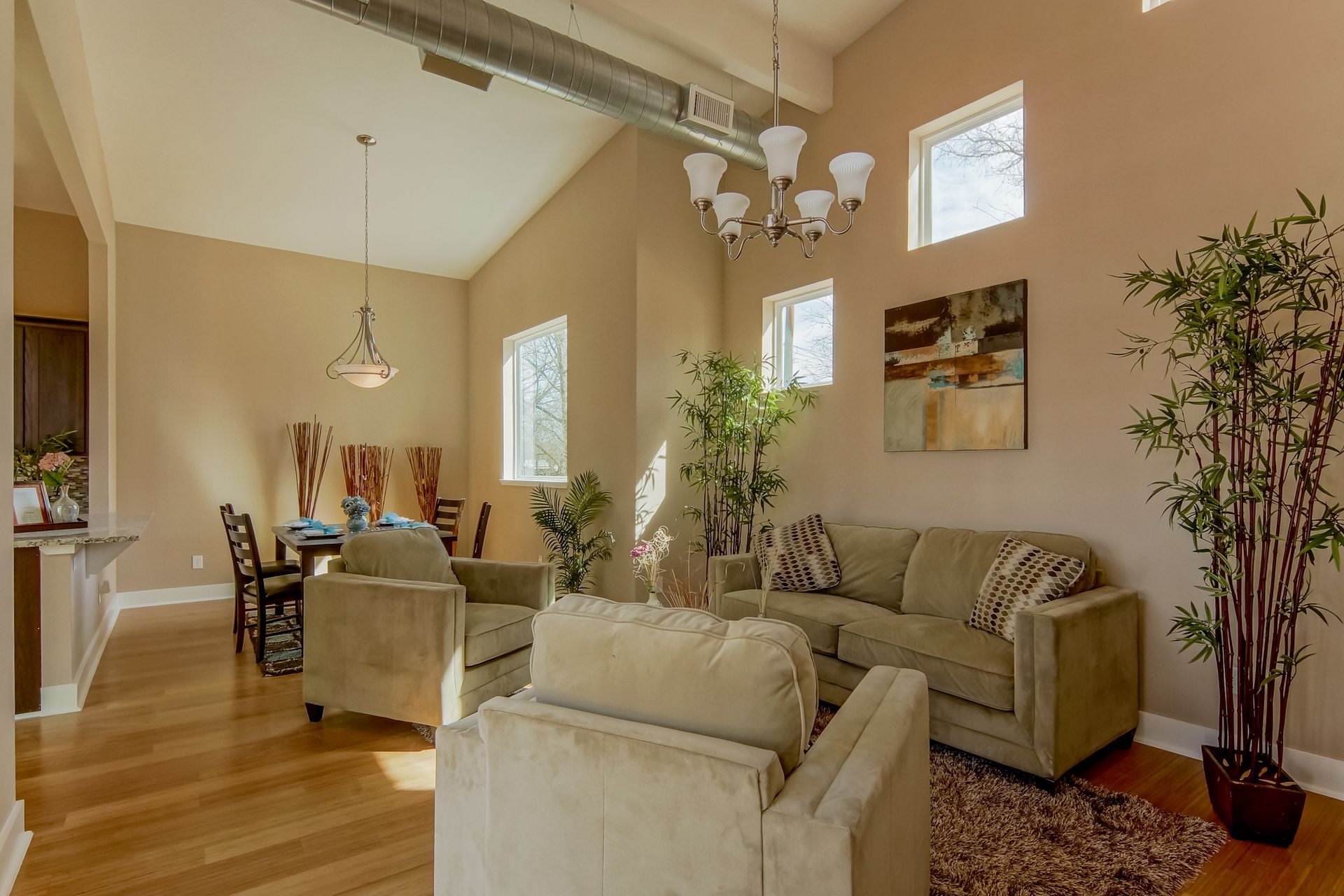 Living room and dining area with neutral furniture, a chandelier, hardwood floors, and potted plants in a bright space.