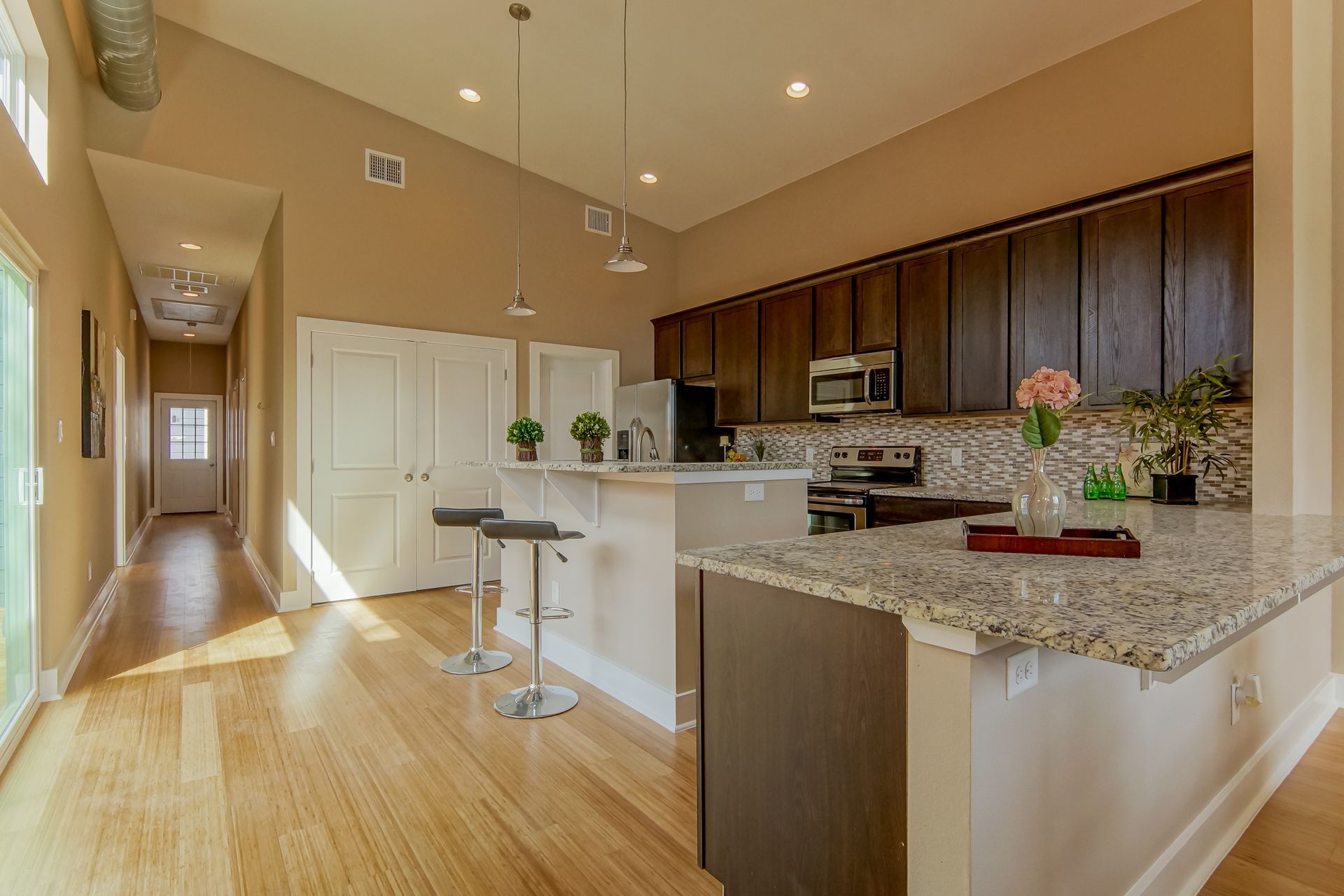 A bright modern kitchen with granite countertops, dark wood cabinets, and bar stools, leading into a sunlit hallway.