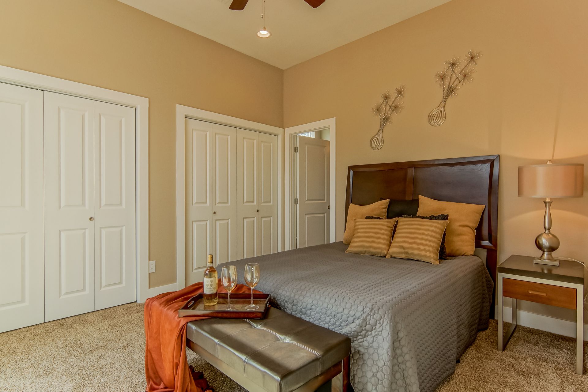 A bedroom with a queen bed, two white closets, a nightstand with a lamp, and a bench with a wine bottle and glasses.