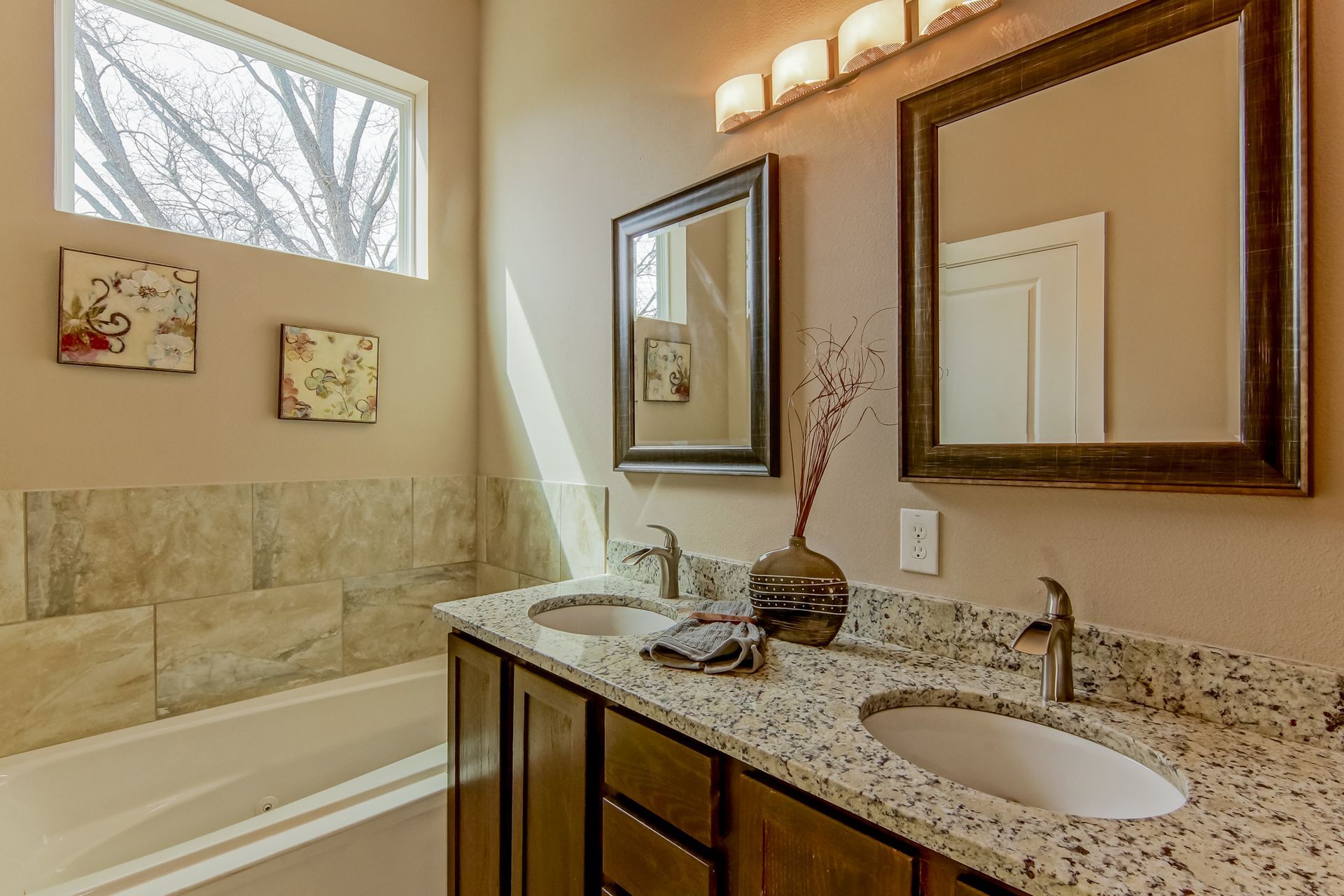 A bathroom with a double sink vanity, granite countertop, two framed mirrors, and a tiled bathtub under a large window.