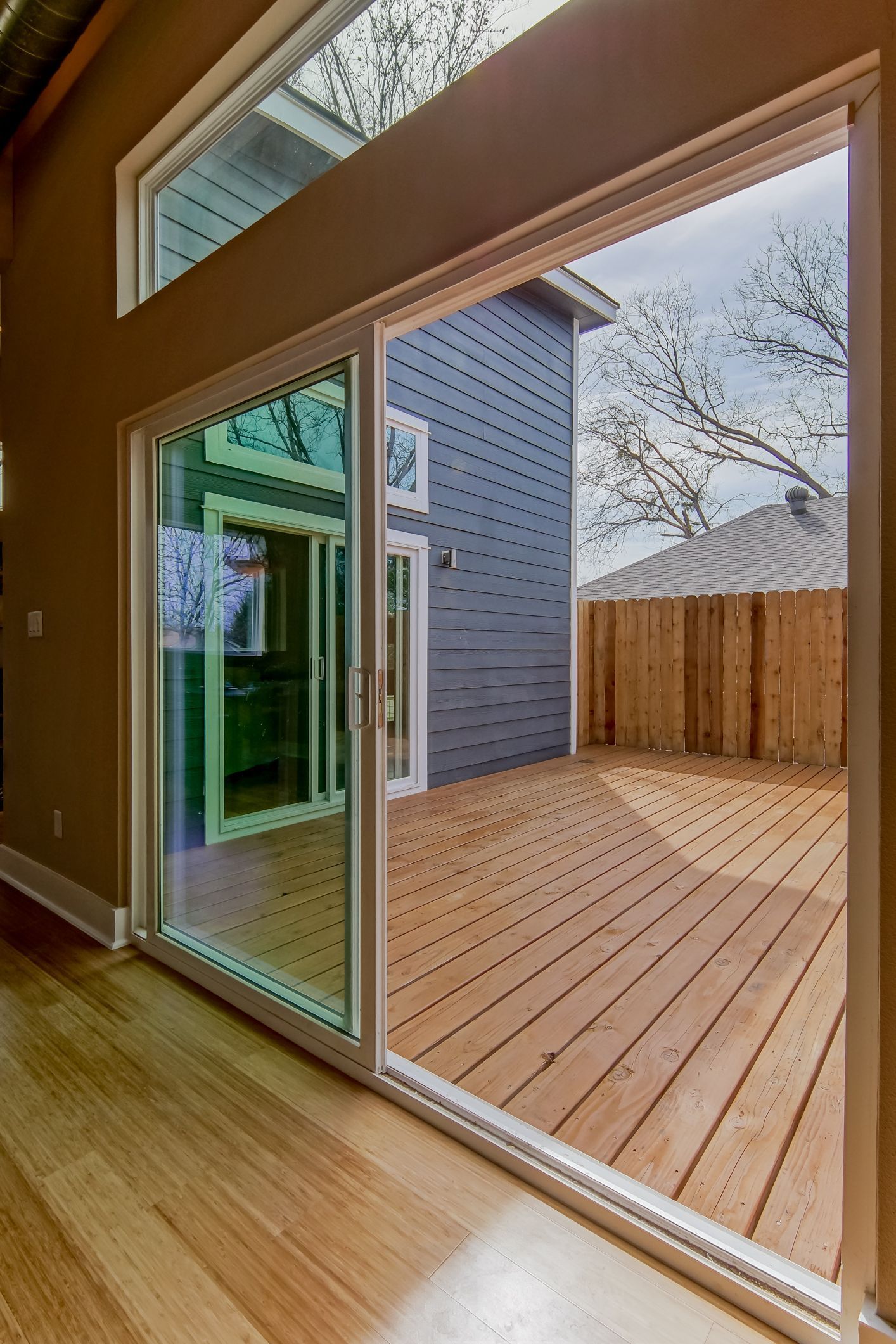 A view from indoors looking through a sliding glass door onto a wooden deck and a house with blue siding.